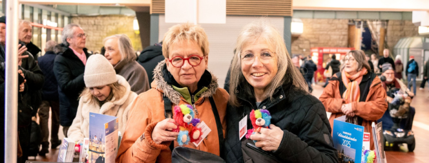 Zwei Frauen mit Teddys im Arm am Teddystand von Hamburg Leuchtfeuer, vor Ihnen aufgereit zahlreiche bunte Teddys.
