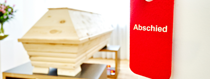 Durch eine geöffnete Tür an der ein roter Anhänger mit dem Wort "Abschied" hängt, blicken wir auf einen Holzsarg und Kerzen in einem hellen Raum.
