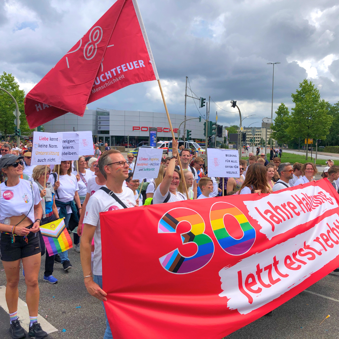 Bei der CSD Demo in Hamburg 2025 sind Mitarbeiter*innen, Ehrenamtliche und Freund*innen von Hamburg Leuchtfeuer zusammengekommen. Sie tragen ein Banner auf dem steht: "30 Jahre Haltung. Jetzt erst recht."